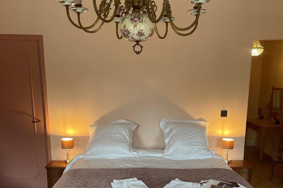 A neatly made bed with a pink quilt, two white pillows, and a pair of towels on the bed. There is a chandelier above the bed, and two bedside lamps are visible.