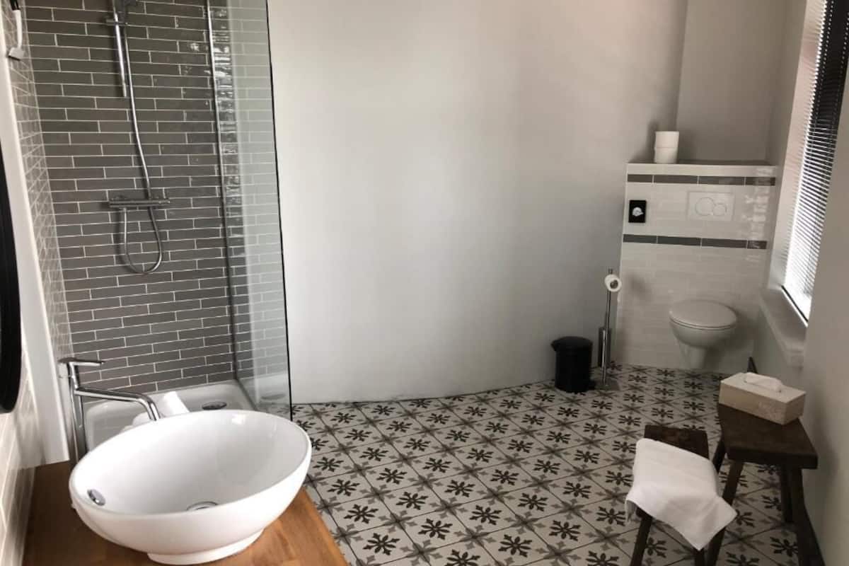 A bathroom with a large white sink, a shower with a glass door, a toilet, and a small table with a towel.