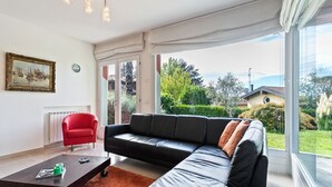 Ein modernes Wohnzimmer mit einer schwarzen Ledersofa, einem roten Sessel und einem Couchtisch. An der Wand hängt ein Gemälde und es gibt einen Blick ins Grüne nach draußen.
