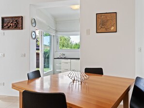 Ruokailutilassa on puinen pöytä, mustat tuolit ja koristeellinen metalliesine. Tilaan kuuluu myös valkoiset kaapit ja ikkuna, josta avautuu näkymä vehreään maisemaan.