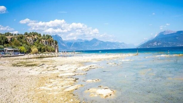 Una playa con agua cristalina, un muelle de madera y montañas al fondo.