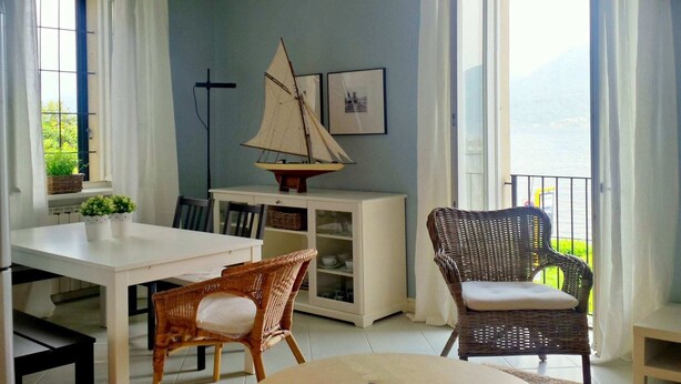 Una habitación con una mesa de comedor, dos sillas de mimbre, un pequeño modelo de velero, una estantería y una ventana con vistas al mar.