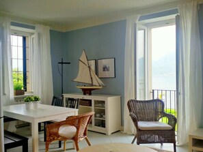Una habitación con una mesa de comedor, dos sillas de mimbre, un pequeño modelo de velero, una estantería y una ventana con vistas al mar.
