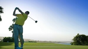 Un golfista en pleno balanceo en un campo de golf con cielo despejado y vista al océano.