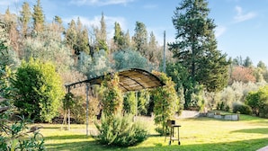 Un jardín con un pérgola, un pequeño carro y diversos arbustos y árboles.