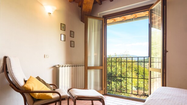 A room with a bed, a chair with a cushion, a radiator, and a view through a large window.