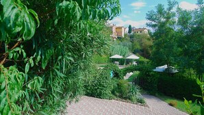 Un sendero de jardín que conduce a un área de descanso con sombrillas, rodeado de abundante vegetación y un edificio distante.