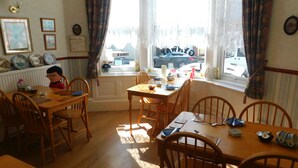 A dining room with a wooden table and chairs, a window with curtains, and a view of parked cars outside. - The Gynway (Blackpool)