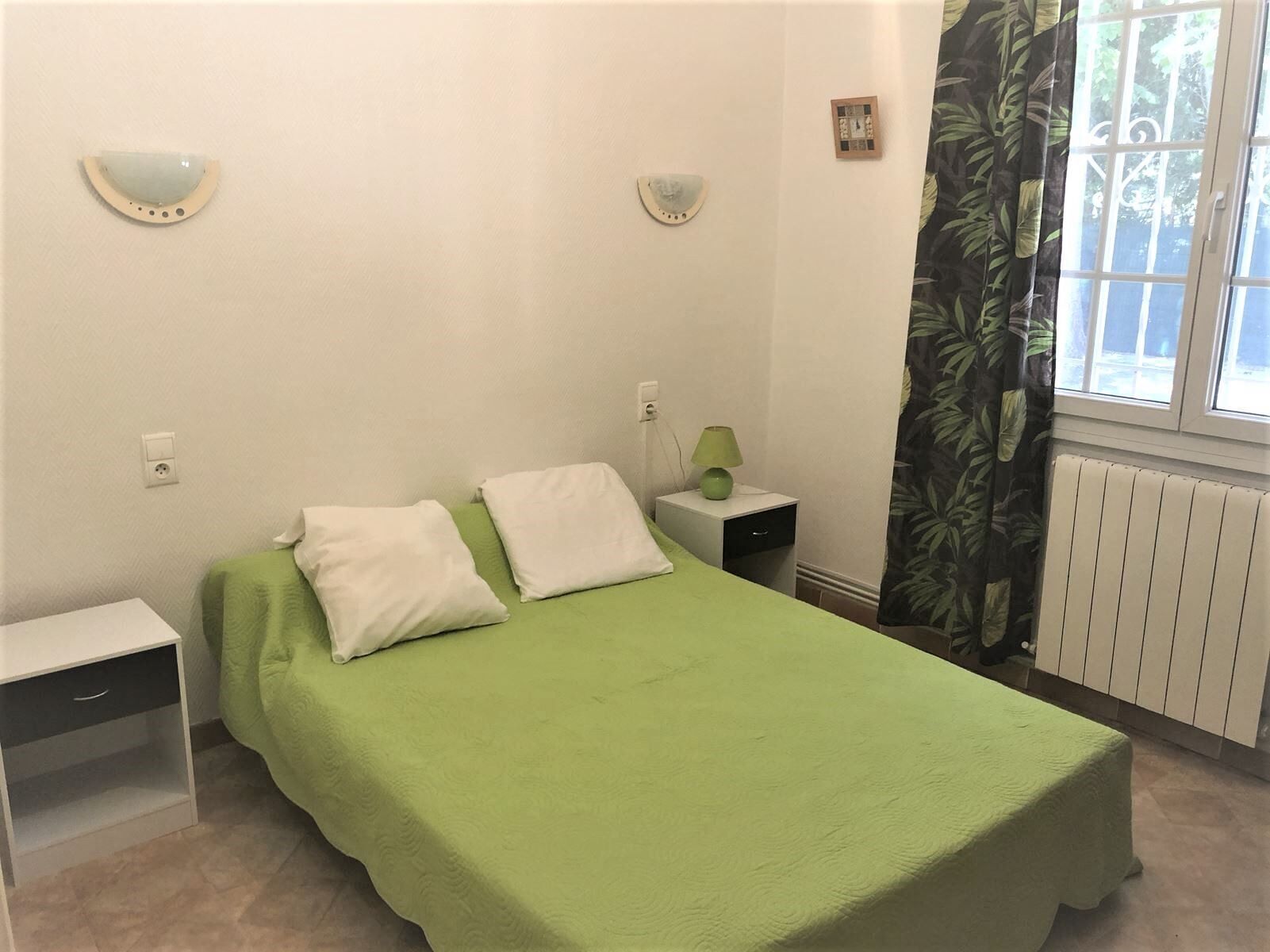 Un dormitori amb un llit individual, dues taules de nit, una estantària amb una làmpada, una finestra amb cortines i un radiador.