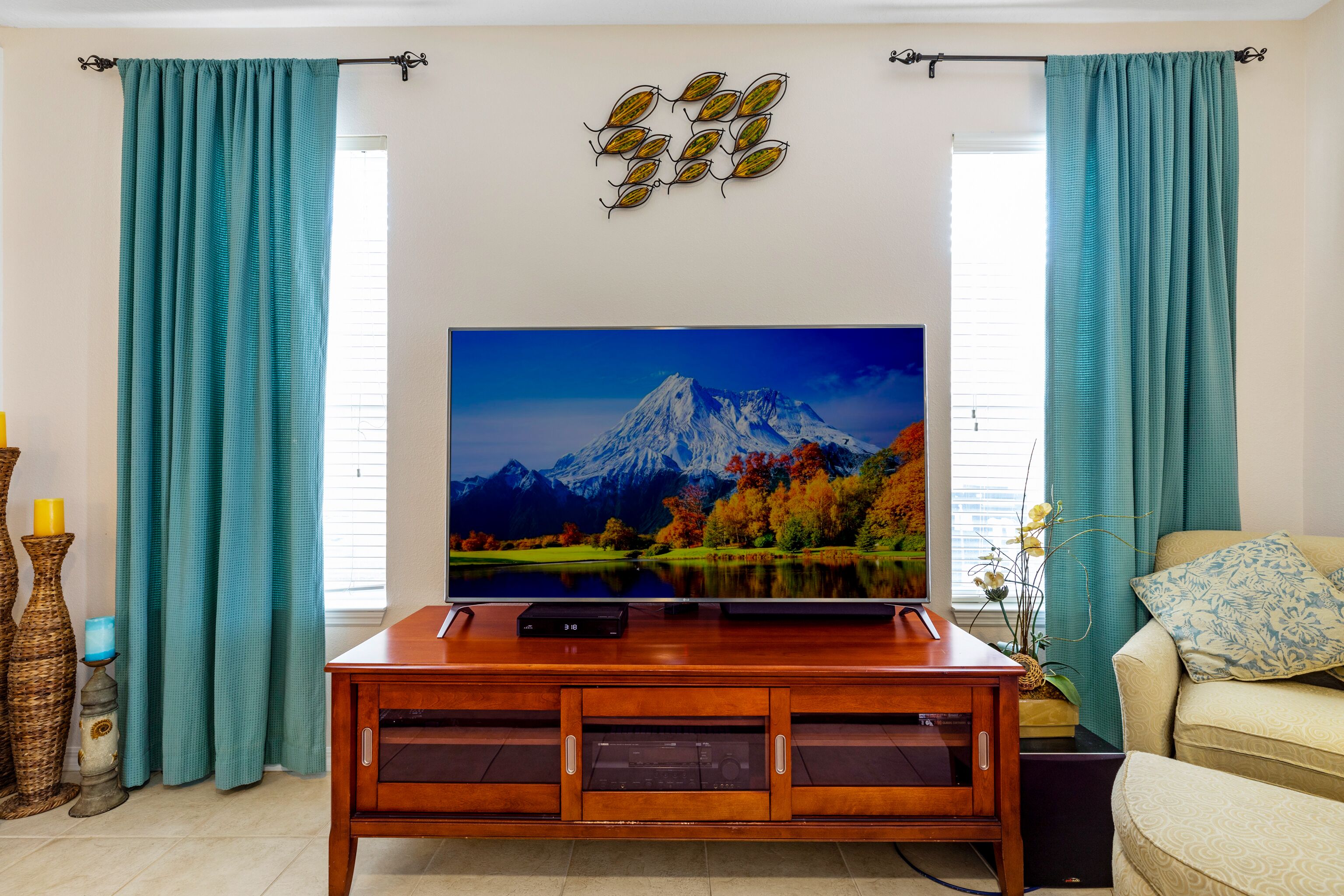 A living room with a large flat-screen TV on a wooden stand, teal curtains, a decorative wall piece, and a beige sofa with a patterned pillow.