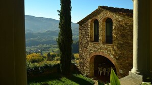 A stone building with an arched entrance and two windows, surrounded by greenery and a mountainous landscape.