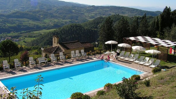 A swimming pool with lounge chairs and umbrellas, surrounded by a scenic landscape with hills and trees.