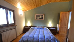 A bedroom with a wooden ceiling, a large bed with a blue quilt, a radiator, a wooden door, and a framed picture on the wall.