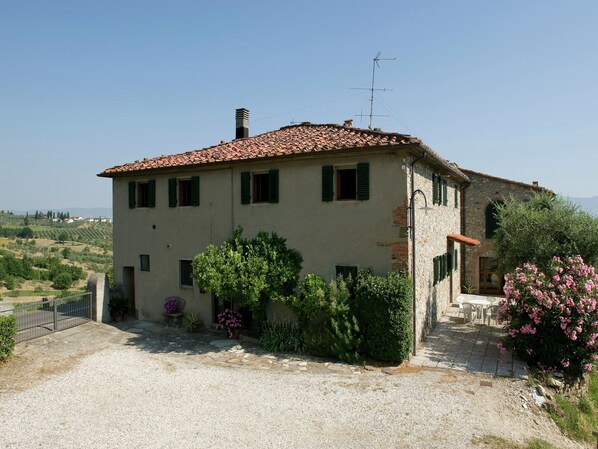 Une maison à deux étages, avec un toit en tuiles, une cheminée et un petit jardin agrémenté de plantes en pot et de fleurs.