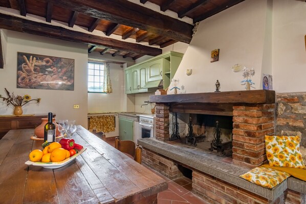 A rustic kitchen with a brick fireplace, wooden table, and a plate of oranges and tomatoes.