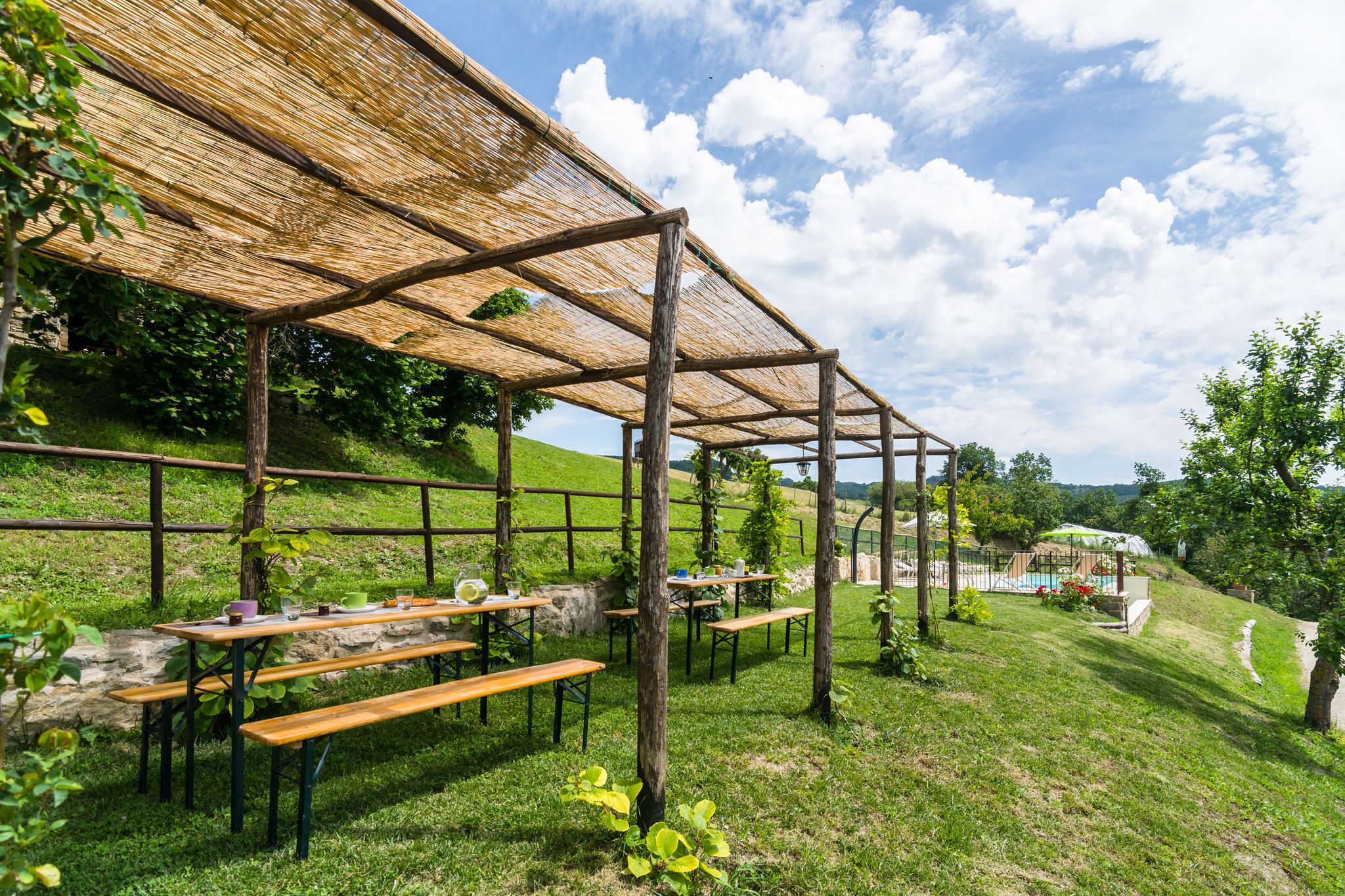 An outdoor seating area with wooden benches and tables under a thatched roof, surrounded by greenery and a stone wall.