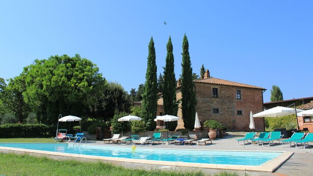 A swimming pool with lounge chairs and umbrellas, surrounded by tall trees and a stone building. - Belvilla by OYO Tiglio (Cortona)