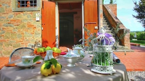 A table set with fruit, teapots, and a birdcage outside a stone building with an open door. - Belvilla by OYO Tiglio (Cortona)