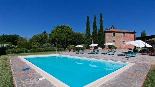 A rectangular swimming pool with a stone border, surrounded by lounge chairs and umbrellas, set against a backdrop of trees and a brick building. - Belvilla by OYO Leccio (Cortona)