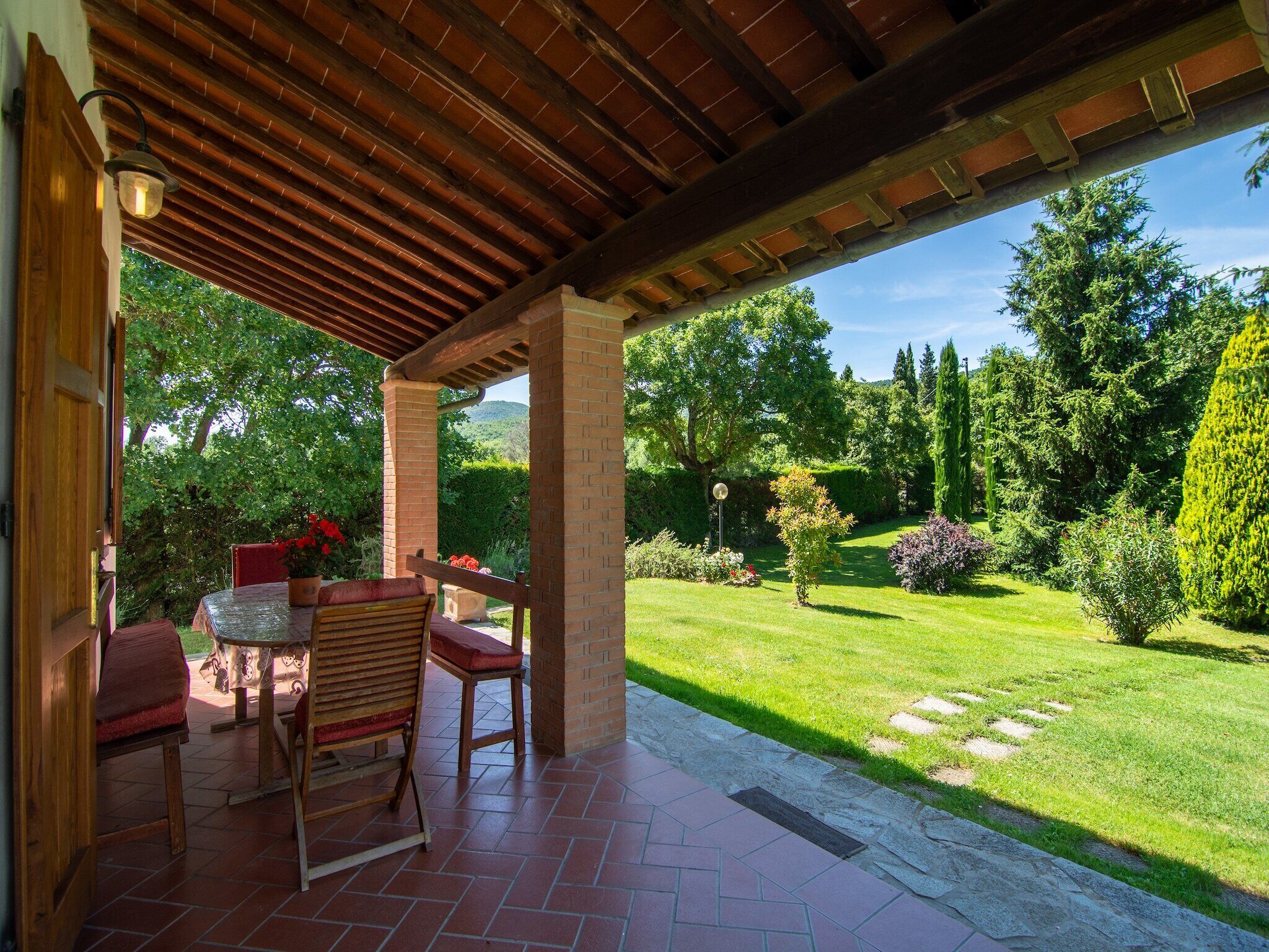 Un patio cubierto con una mesa y sillas, rodeado de un jardín cuidado y vistas al campo.