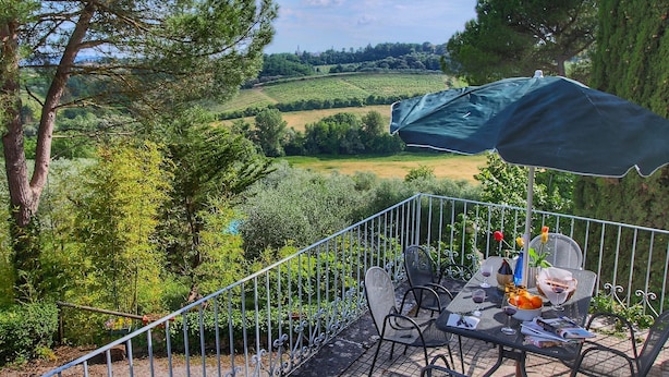 A terrace with a table set for dining, surrounded by a metal railing, overlooking a scenic landscape with trees and fields. - Belvilla by OYO Lunghe (Siena)