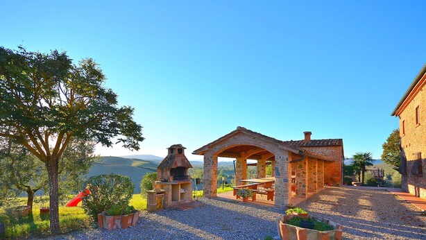 Un patio in pietra con un'area barbecue in mattoni, circondato da piante in vaso e vista su colline lontane.