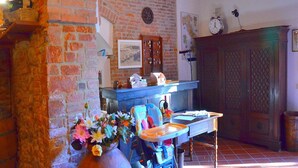 Una stanza rustica con pareti in mattoni a vista, soffitto in legno e pavimento piastrellato. Si nota un tavolo azzurro con una sedia, un vaso grande con fiori e un armadio in legno con una sveglia appesa al muro.