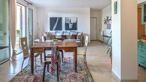 Ein modernes Wohnzimmer mit einem Holzesstisch, Stühlen und einer Couch. Ein großes Fenster mit Vorhängen, eine Deckenleuchte und eine grüne Kommode mit Schubladen sind ebenfalls zu sehen.