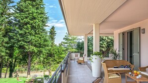 Ein überdachter Balkon mit Gartenmöbeln, Blick auf eine grüne Landschaft und ein Gebäude mit Rollläden.