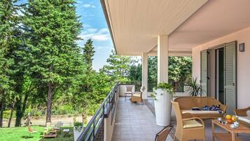 Een balkon met rieten meubels, een tafel met fruit en uitzicht op een groene tuin.