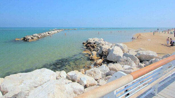 Ein Strand mit klarem Wasser, felsigem Ufer und einer Holzpromenade.
