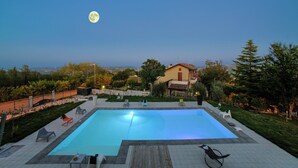 Ein Schwimmbad mit Holzterrasse, umgeben von Sitzmöbeln im Freien und Blick auf ein Haus und die Landschaft.