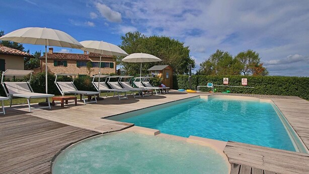 A swimming pool with sun loungers and umbrellas, surrounded by a wooden deck and a building with a tiled roof. - Cottage in Montelabbate with Pool & Forest (Montelabbate)