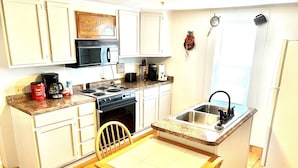 A compact kitchen with white cabinets, a stove, a microwave, a sink, and a dining area with a table and chair.
