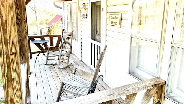 A wooden porch with a rocking chair and a table, a sign reading "Chandler Home", and a window with blinds.