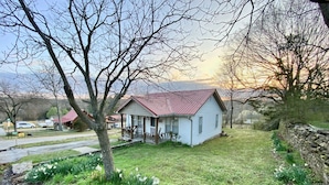 A small white house with a red roof, surrounded by a grassy yard and flowering plants.