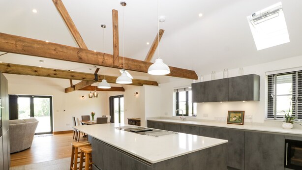 A modern kitchen with a large island, wooden beams, pendant lights, and a view of the outdoors. - The Old Dairy (Wokingham)