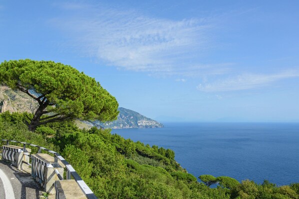 A coastal road with a guardrail, lush greenery, and a clear view of the sea and distant land.