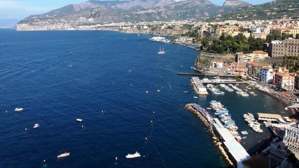 A coastal city with a marina, boats, and a mountainous backdrop.