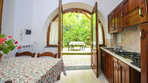 A kitchen with wooden cabinets, a dining table with a floral tablecloth, and a view of a garden through an open doorway.