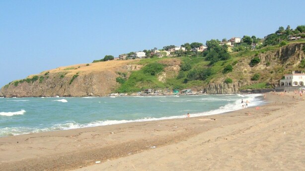 A coastal beach with a rocky cliff and houses on top.