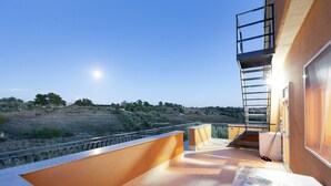 A balcony with a view of a hilly landscape, a metal staircase, and a covered area with a table and chairs.