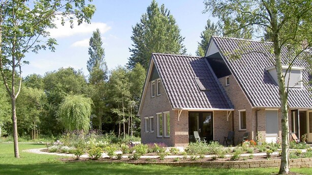 A two-story house with a sloped roof, brick exterior, and a landscaped garden with a lawn and trees. - Rural holiday home with terrace (Zuidwolde)