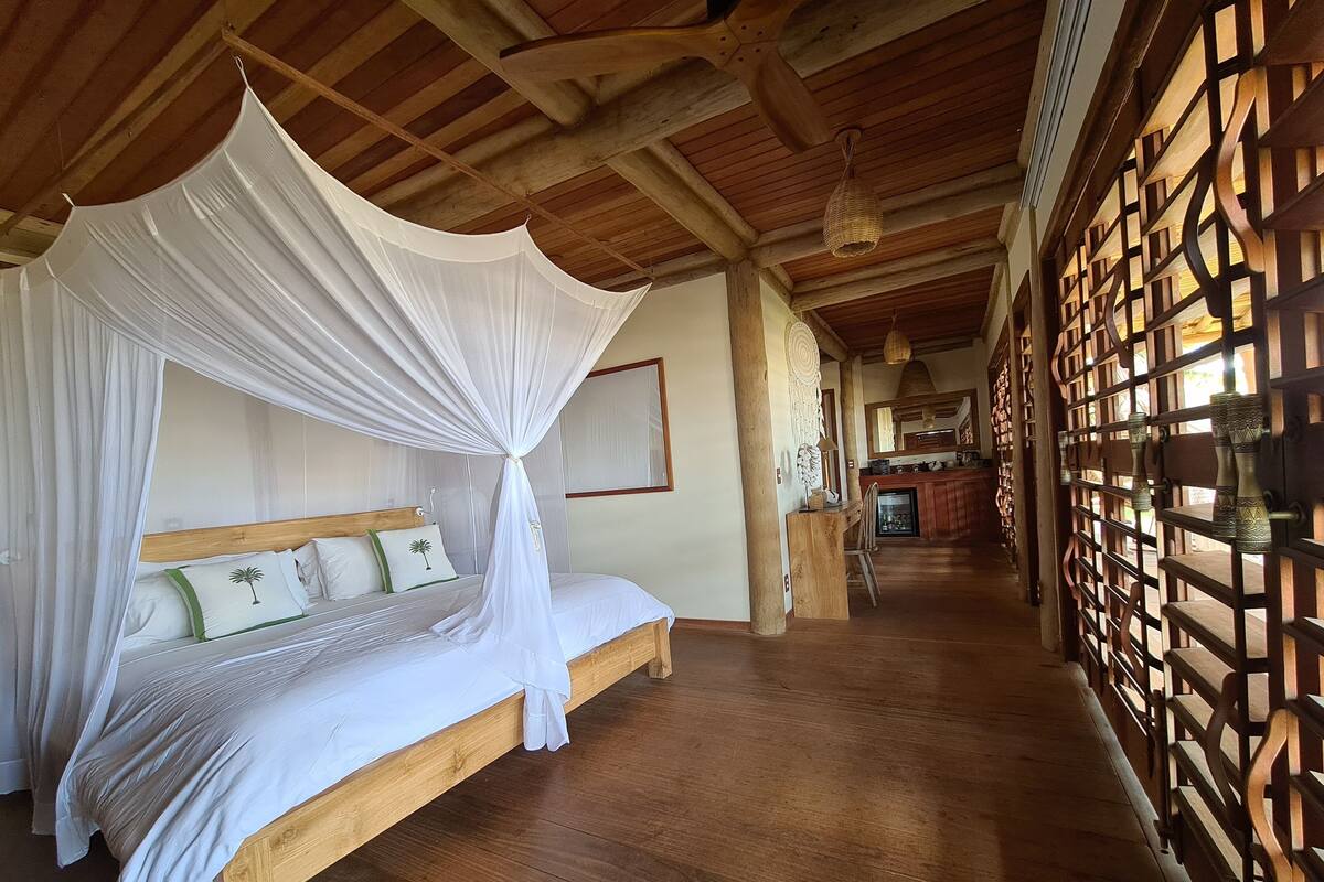 Una habitación con cama bajo un dosel, vigas de madera en el techo, una cocina al fondo y una pared decorativa con diseño de celosía.