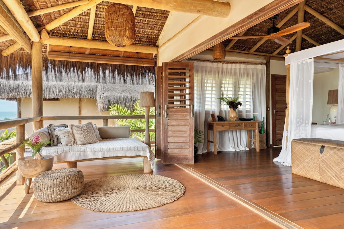 Una terraza de madera con un sofá, un reposapiés, un tapete tejido y una mesa de madera con una lámpara.