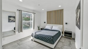 Ein modernes Schlafzimmer mit Bett, Nachttischen, einem Fenster mit Vorhängen und Kunstwerken an den Wänden.