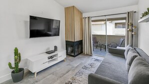 Ein modernes Wohnzimmer mit einem Flachbildfernseher, einer weißen Unterhaltungsanlage, einem grauen Sofa, einem gemusterten Teppich und einer Kakteenpflanze.