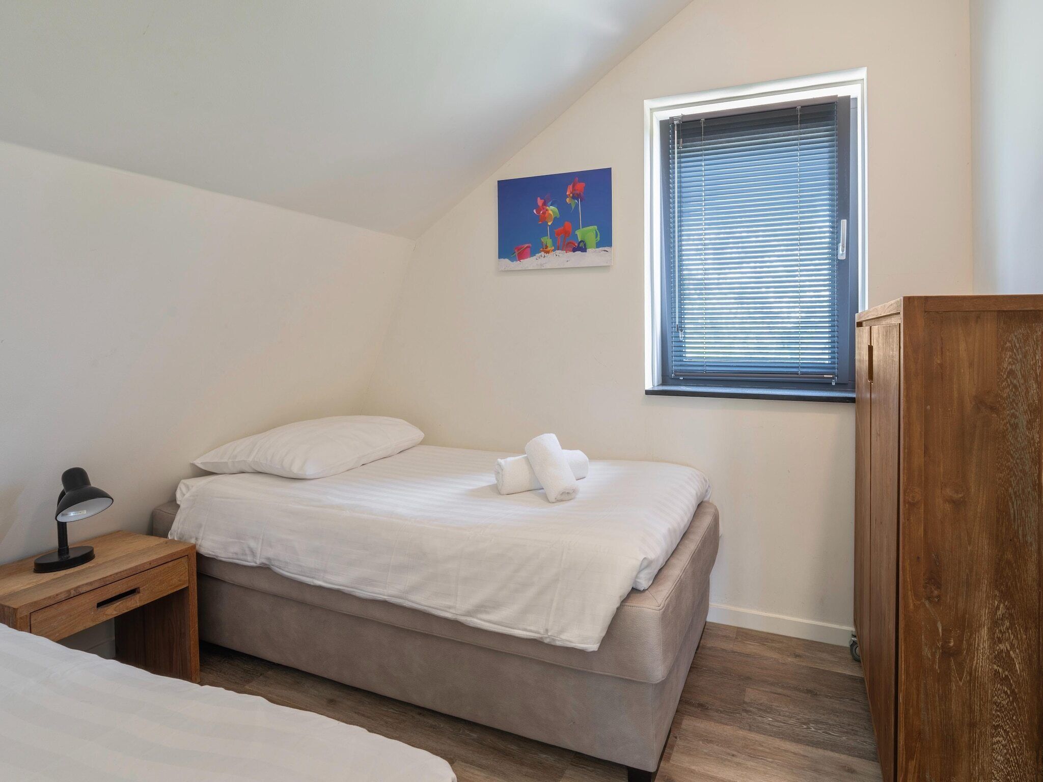 A bedroom with a single bed, a nightstand with a lamp, a wooden wardrobe, a window with blinds, and a colorful painting on the wall.