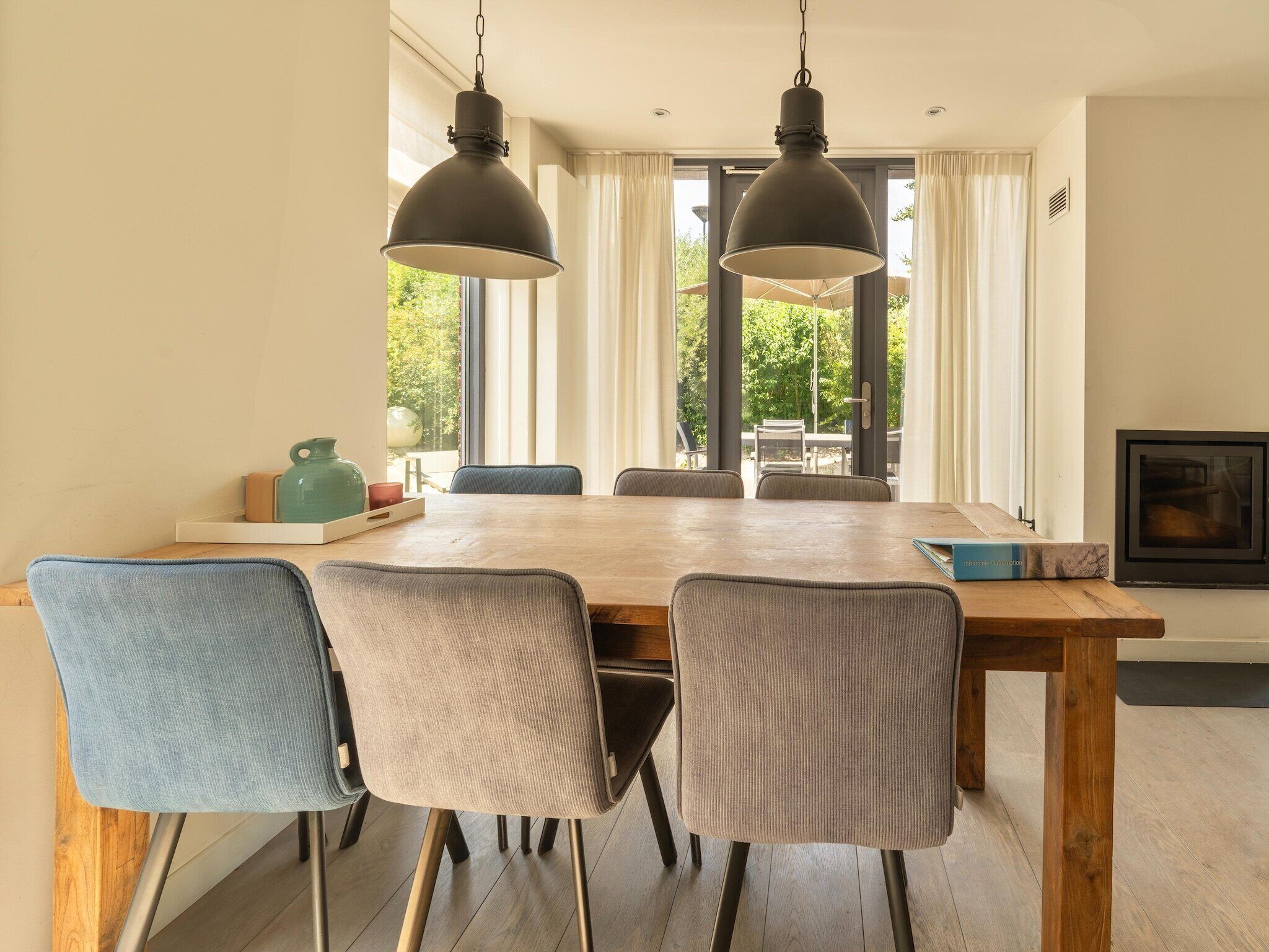 Comedor con mesa de madera, cuatro sillas, dos lámparas colgantes y vista a zonas verdes al exterior.