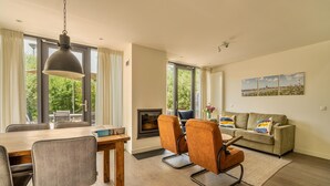 A modern living room with a fireplace, a dining area with a wooden table and chairs, and large windows offering a view of greenery.